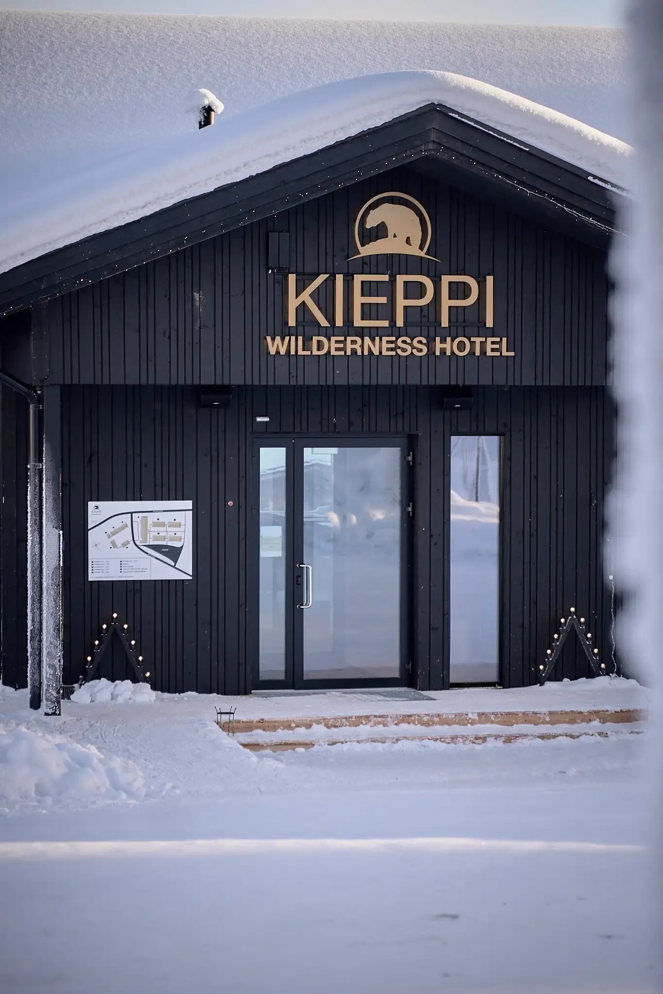 Hirsirakennus lumisessa maisemassa. Oven yläpuolella teksti: Kieppi Wilderness Hotel.