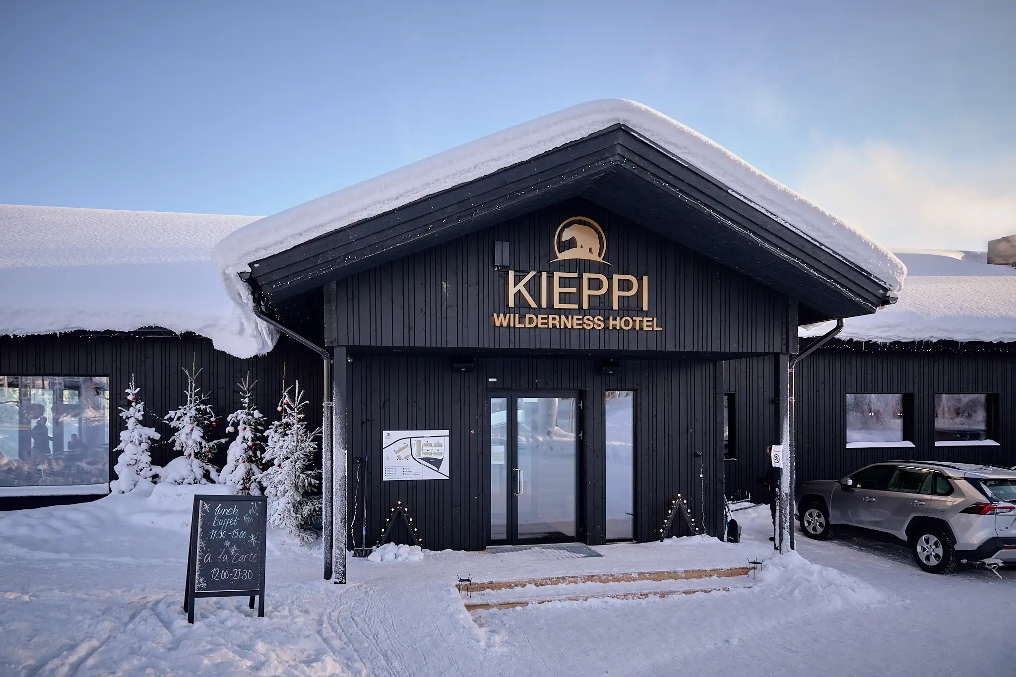 Hirsirakennus lumisessa maisemassa. Oven yläpuolella teksti: Kieppi Wilderness Hotel.