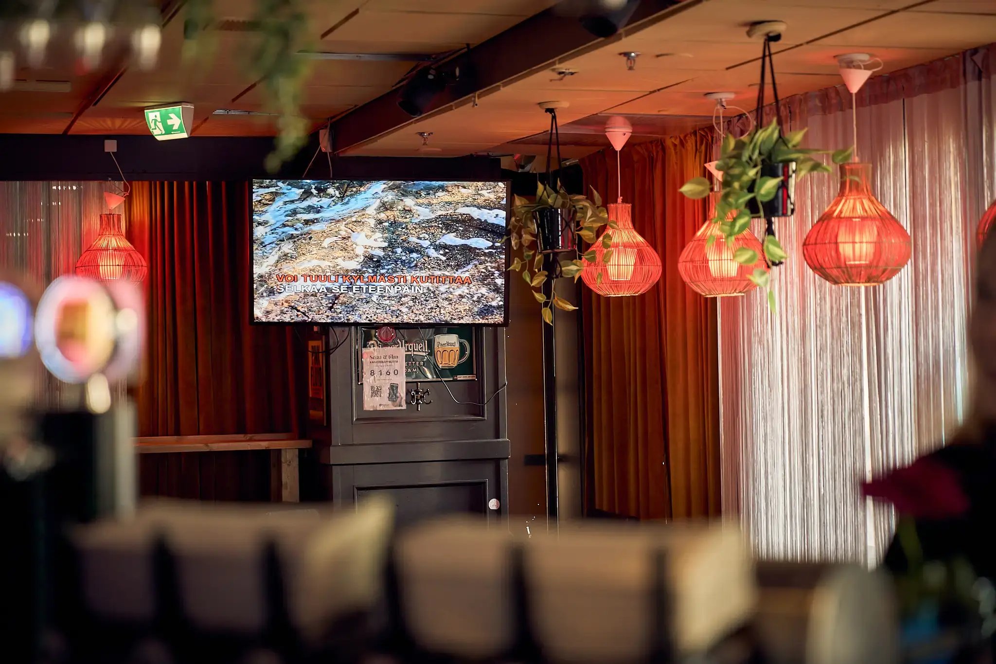 Ravintolan seinällä oleva televisioruutu, jossa pyörii karaokekappale. Vieressä punaisia lamppuja.
