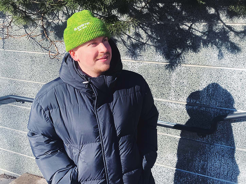 Young man wearing a puffy winter jacket and a green beanie. He is outside in sunny winter weather. 