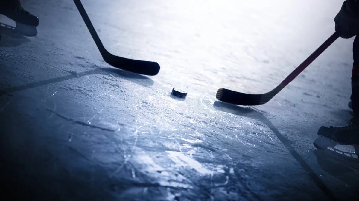 Two hockey sticks and a hockey puck on the ice under dim lighting.