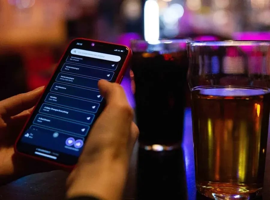 Ihmisen käsissä oleva puhelin, jossa näkyy listaus karaokebiiseistä. Vieressä kaksi tuoppia.