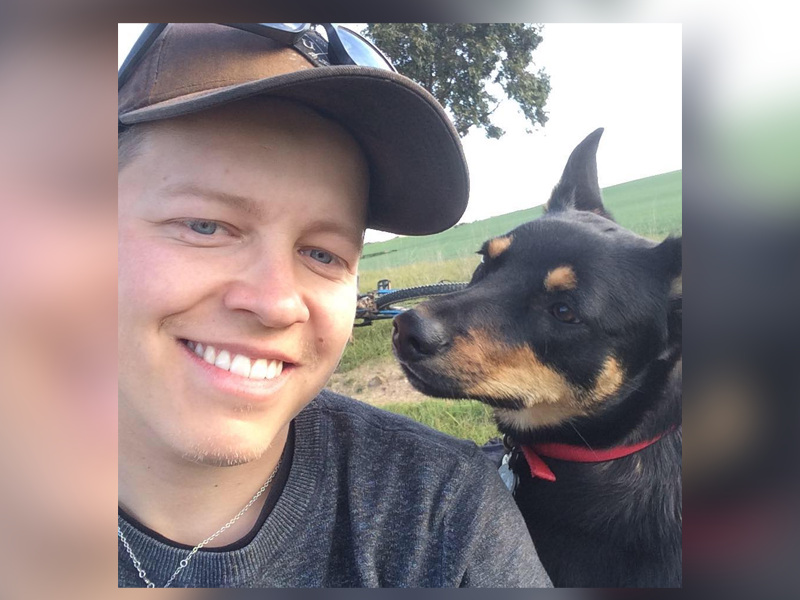 A smiling young man on some grass wearing a cap. There is also dog next to him.