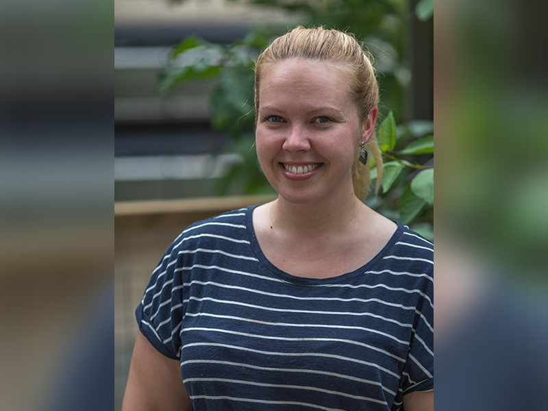 Smiling young woman wearing a stripe shirt outside in front of a leafy tree.