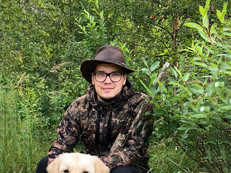 Young man wearing hunting gear in a forest. Also a glimpse of a dog at the bottom half of the image.