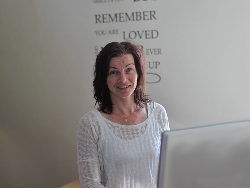 Woman wearing a white shirt, looking into the camera while sitting down in front of a computer.