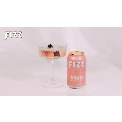 Fizz Spritz -tölkki vaaleanpunaisella taustalla. Vieressä lasi, jossa juomaa.