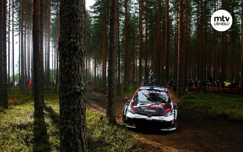 Ralliauto metsän keskellä. Taustalla Suomen lippuja.
