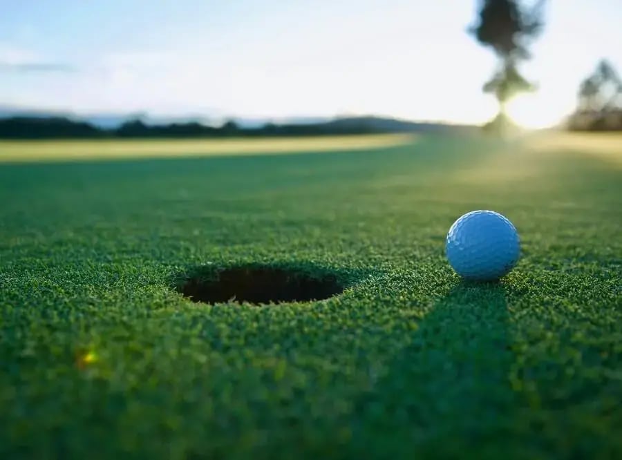 A golf ball on the green, close to the hole, with the sun shining in the background.