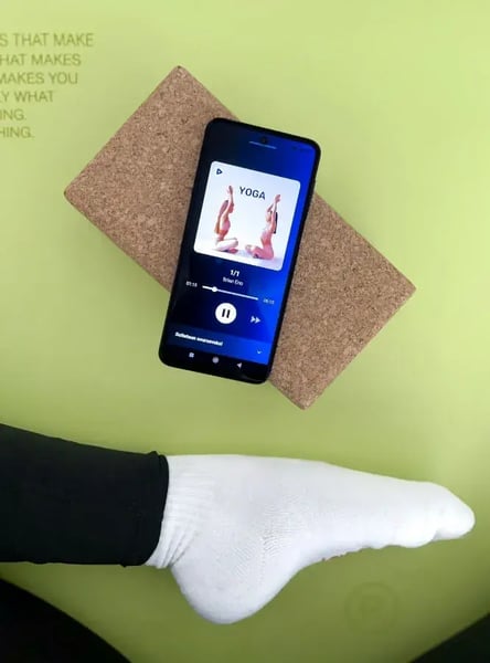 A green yoga mat with a wooden block and a phone on top. The phone displays the Feelment app.