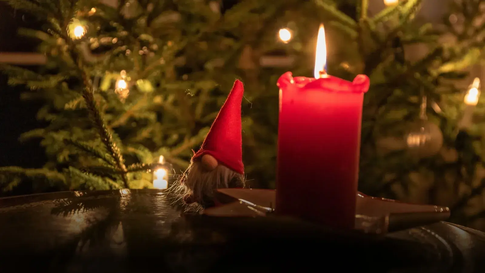 Punainen kynttilä ja tonttu-ukko pöydällä. Taustalla joulukuusi valoineen.