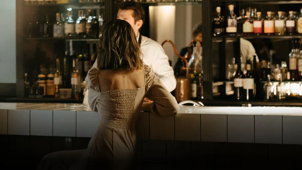 A woman in a light-colored dress leaning on the bar counter, captured from behind. Behind the woman, a bartender is preparing a drink.