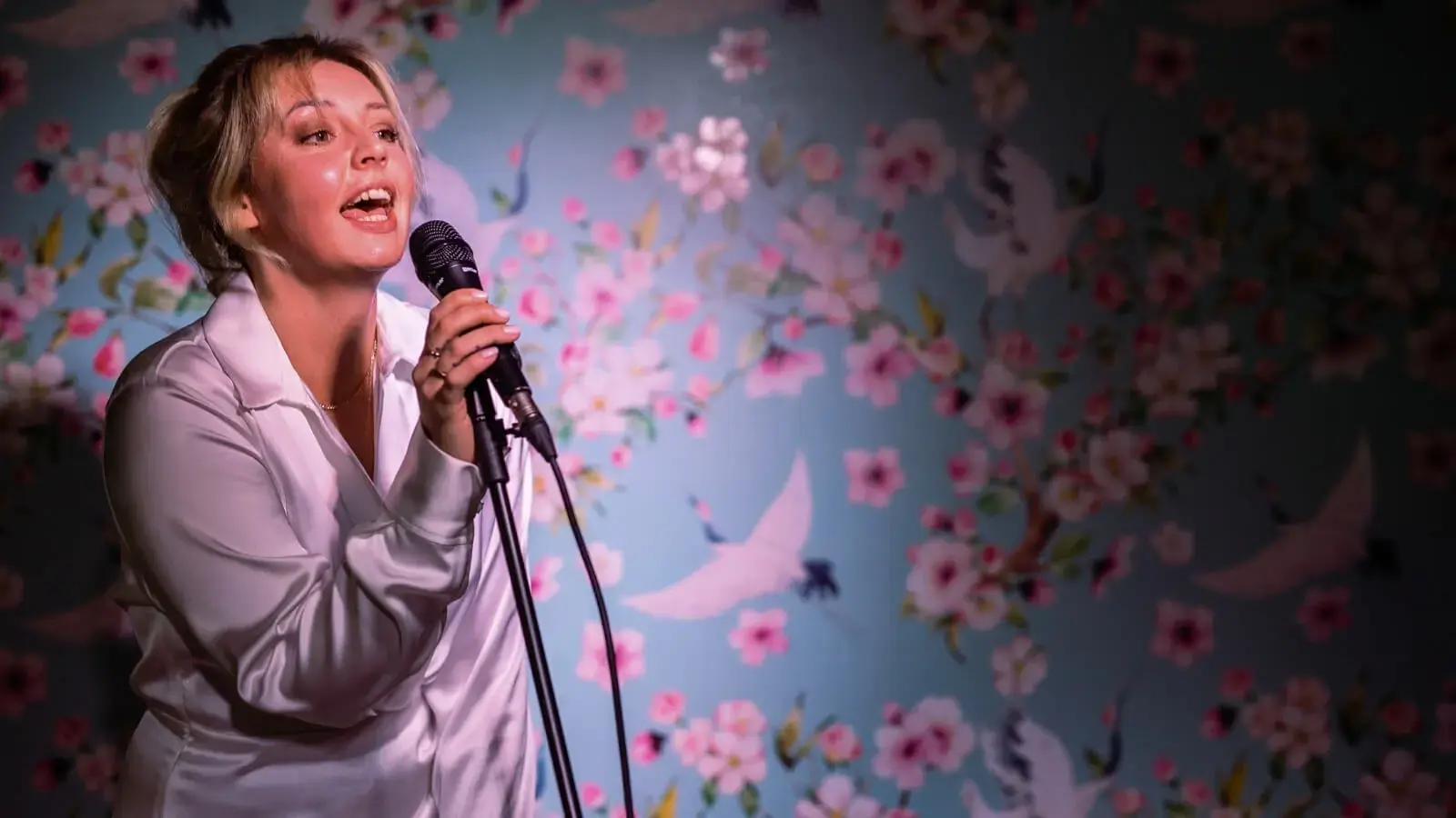 Nainen laulamassa karaokea. Taustalla seinä, jossa vaaleanpunaisia kukkia.